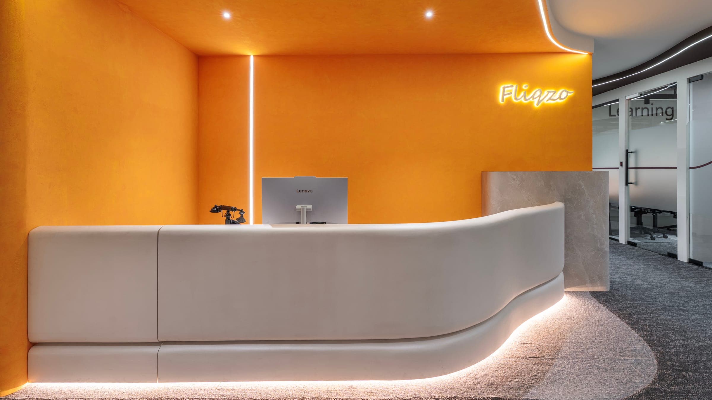 Fliqzo reception with a backlit logo on a warm orange feature wall and sculptural white desk, Coimbatore